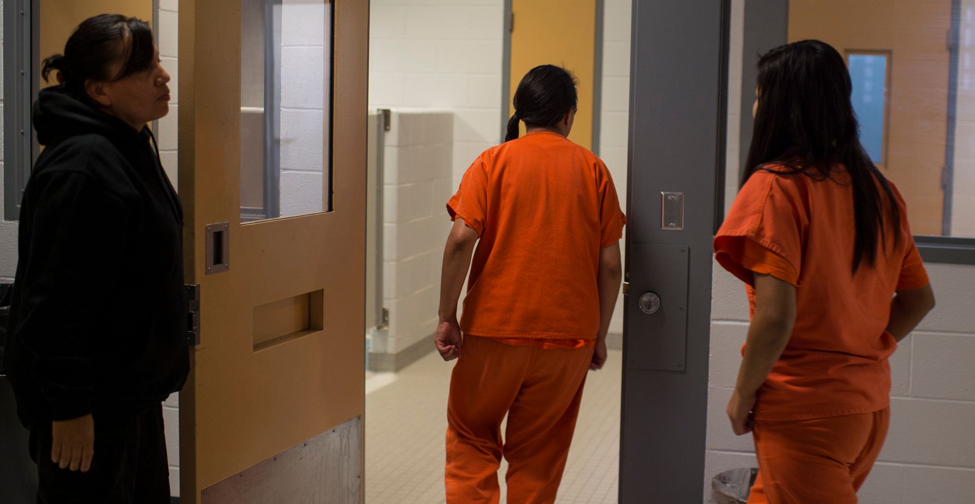 Two young people with long brown hair, wearing orange uniforms, walk toward an open door as an adult wearing a black sweatshirt holds the door open.   