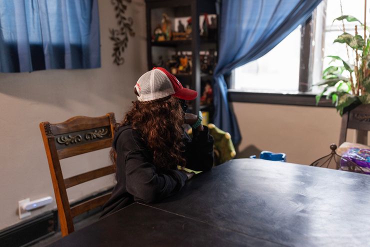 A woman with long, wavy red hair, wearing a baseball cap and whose face is not visible, sits at a table looking out a window with blue curtains. 