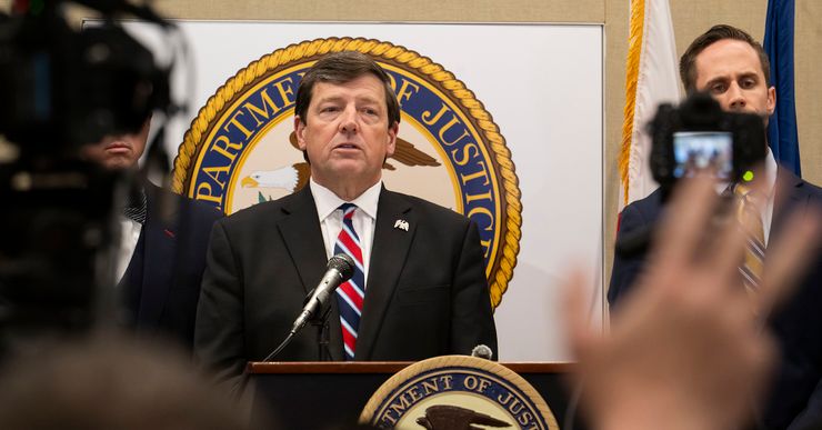 A White man, wearing a black suit and a blue and red tie, talks into a mic while standing at a podium with a U.S. Department of Justice seal on it. A group of reporters holding cameras and microphones stand in front of him. 