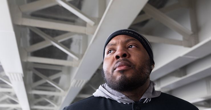 A photo of a Black man with a beard looking to the side under a concrete bridge. The man is wearing a black beanie and jacket and a gray hoodie. 